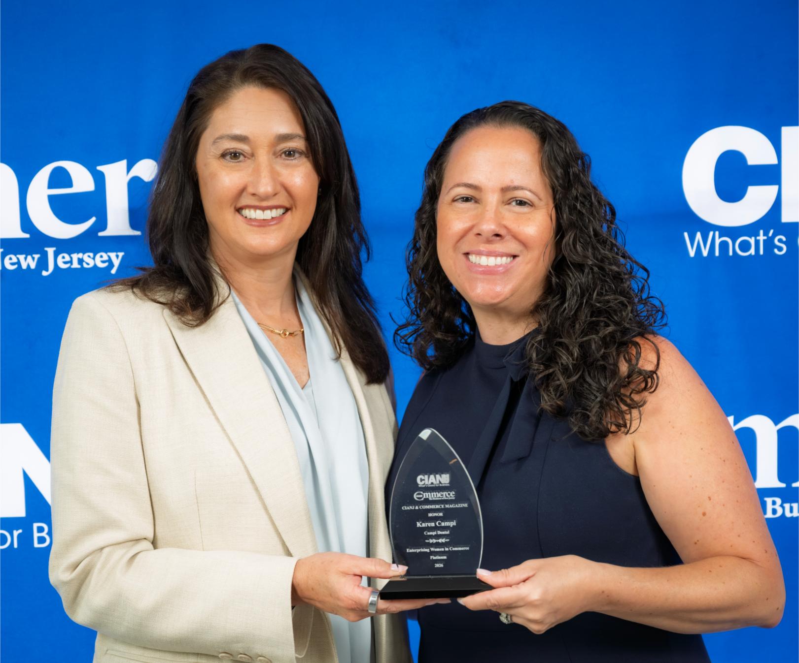 Two professional women smiling while holding a CIANJ Enterprising Women in Commerce award trophy in front of a blue backdrop.